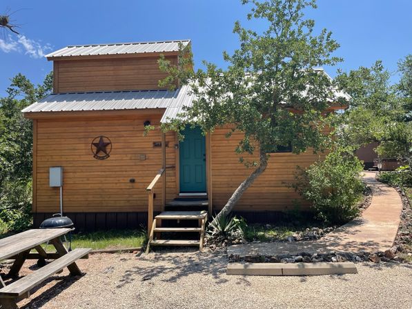 Rustic tiny wooden cabin with metal roof and teal front door, front steps and star wall ornament, picnic table and grill on gravel, shaded by a leaning oak and a winding stone path.