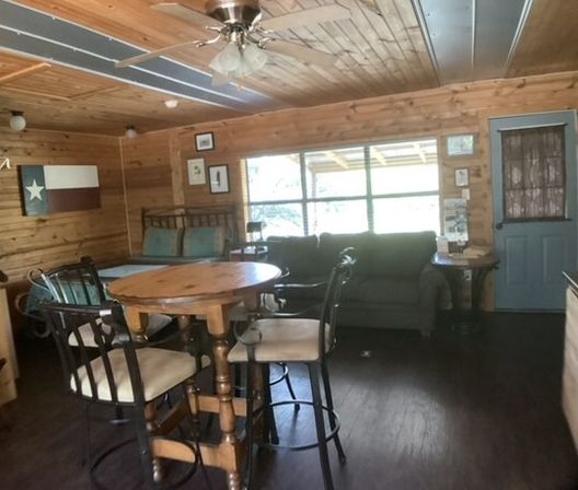 Cozy rustic wood-paneled cabin interior with a round farmhouse table and tall chairs, green sofa beneath a large window, ceiling fan and blue painted door.