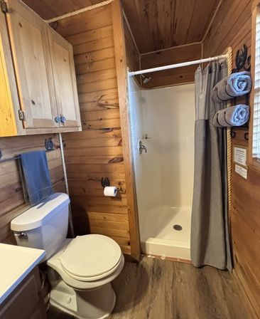 Cozy rustic cabin bathroom with wood-paneled walls, white toilet, compact walk-in shower with gray curtain, rolled gray towels on a wall rack and a wooden storage cabinet.
