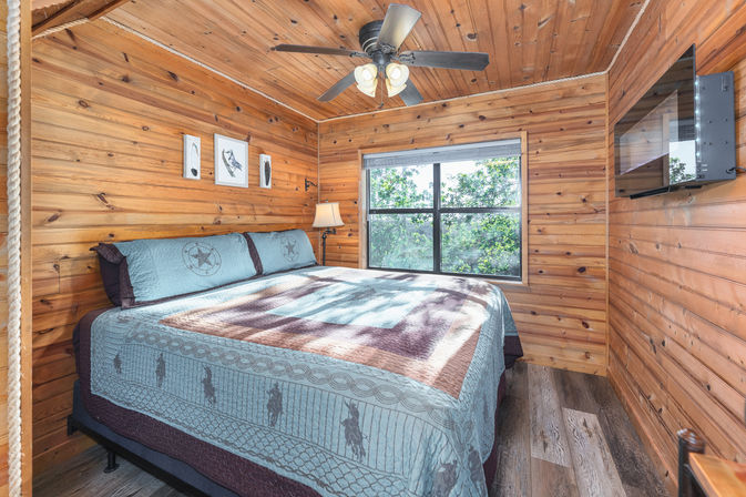 Sunlit rustic cabin bedroom with wood-paneled walls and ceiling, king bed with patterned blue quilt, ceiling fan, bedside lamp, wall art, wall-mounted TV and large window overlooking leafy trees.