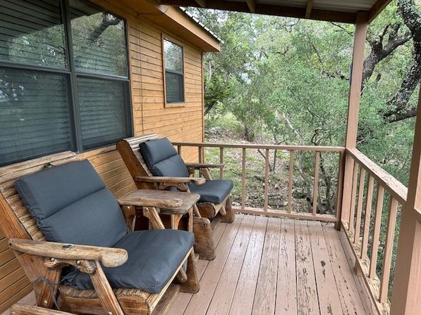 Inviting rustic cabin porch with two cushioned log armchairs and a small wooden side table on a weathered deck overlooking a wooded setting