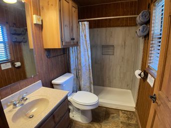Cozy rustic cabin-style bathroom with wood-paneled walls, vanity sink, toilet, walk-in shower with floral curtain and rolled towels by a window