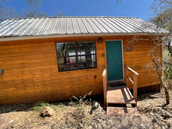 Rustic wooden cabin with teal front door, metal roof and small steps, surrounded by dry rocky landscaping and trees under a clear blue sky