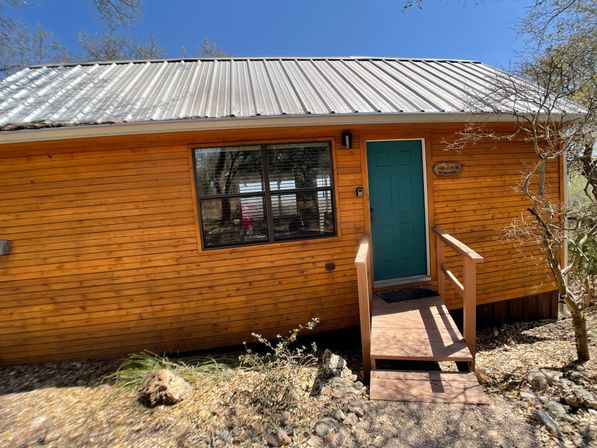 Rustic wooden cabin with teal front door, metal roof and small steps, surrounded by dry rocky landscaping and trees under a clear blue sky