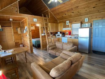 Cozy wood‑paneled cabin interior showing open-plan living area and kitchenette with two tan sofas, small dining table, stainless refrigerator, stove and microwave, ladder to a loft and a visible bedroom doorway.