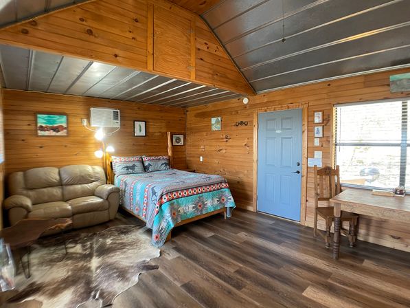Cozy rustic cabin bedroom interior with wood-paneled walls and metal ceiling, queen bed with colorful patterned quilt, leather loveseat, cowhide rug, blue entry door, wooden table and chair by window, wall air conditioner and wood-look floors.