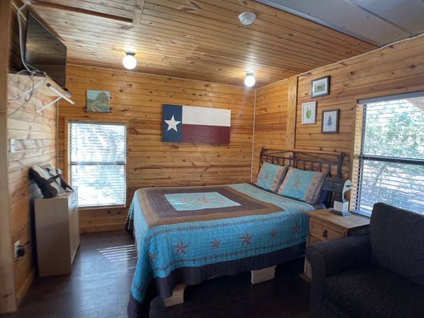 Cozy rustic wood‑paneled cabin bedroom with a turquoise star‑pattern quilt on a queen bed, Texas flag wall art, bedside table, armchair, two windows with blinds and a wall‑mounted TV.