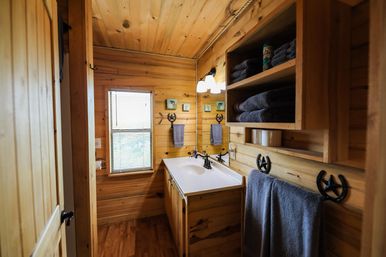 Cozy rustic cabin bathroom interior with knotty pine walls and ceiling, white vanity sink with bronze faucet, mirror and sconce, open shelves of folded gray towels, horseshoe towel hooks, and a window with blinds
