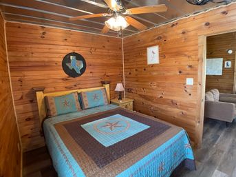 Cozy rustic wood-paneled cabin bedroom with a star-pattern turquoise and brown quilt on a wooden bed, bedside lamp, ceiling fan, and Texas-themed wall art.