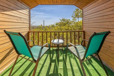 Sunny wooden cabin balcony with two teal sling chairs, a small round table on green turf, and treetop views under a clear blue sky.