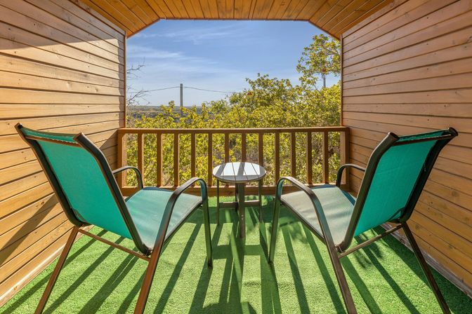 Sunny wooden cabin balcony with two teal sling chairs, a small round table on green turf, and treetop views under a clear blue sky.