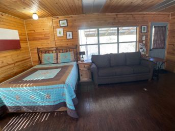 Cozy rustic wood‑paneled cabin room with queen bed in a turquoise star quilt, wooden nightstand and lamp, gray sofa by a large sunlit window, and dark hardwood floor.