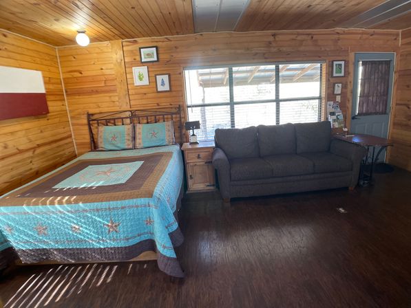 Cozy rustic wood‑paneled cabin room with queen bed in a turquoise star quilt, wooden nightstand and lamp, gray sofa by a large sunlit window, and dark hardwood floor.