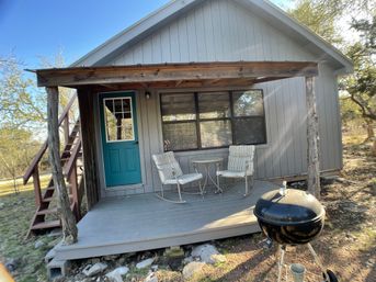 Cozy gray cabin porch in a wooded rural setting with a teal door, two striped rocking chairs and round table under a rustic wood awning, black kettle charcoal grill in the foreground.