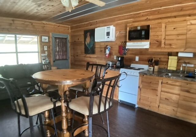 Cozy rustic cabin interior with knotty pine walls and ceiling, round wooden dining table with four cushioned metal chairs, small kitchenette with white stove, microwave, sink and coffee maker, wall air conditioner, sofa by a large window, ceiling fan and rooster wall art.