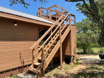 Sunlit wooden staircase leading to a rooftop viewing deck on a brown cabin, surrounded by trees and a gravel path under a clear blue sky