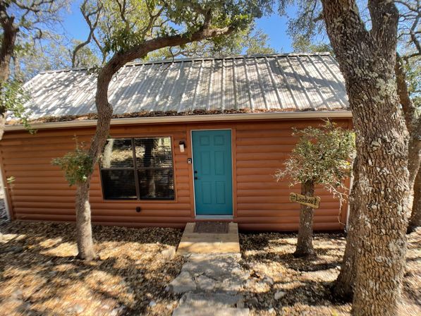 Cozy rustic log cabin with a teal front door and corrugated metal roof, framed by oak trees and a stone pathway in a wooded rural setting.