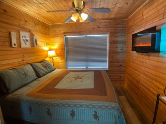 Cozy rustic cabin bedroom with wood-paneled walls and ceiling, king bed with star-patterned quilt, ceiling fan, bedside lamp by a window with blinds, wall-mounted electric fireplace and bird art.