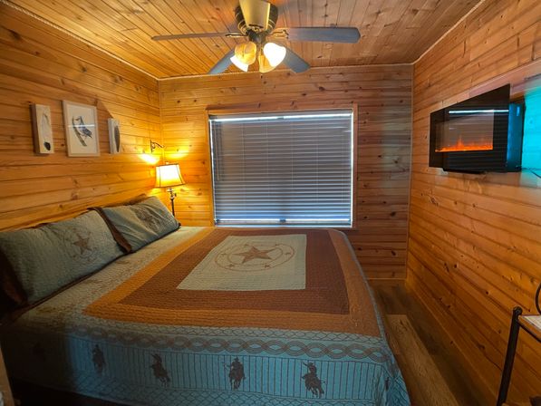 Cozy rustic cabin bedroom with wood-paneled walls and ceiling, king bed with star-patterned quilt, ceiling fan, bedside lamp by a window with blinds, wall-mounted electric fireplace and bird art.