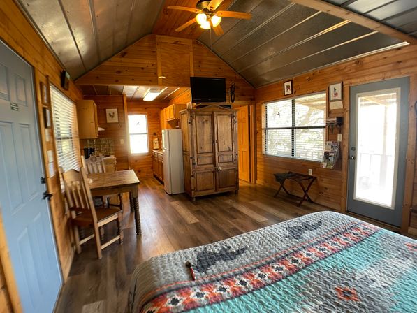 Cozy rustic cabin studio interior with wood-paneled walls, colorful quilted bed, kitchenette, dining table, wooden armoire, ceiling fan, large window and glass door to a porch.