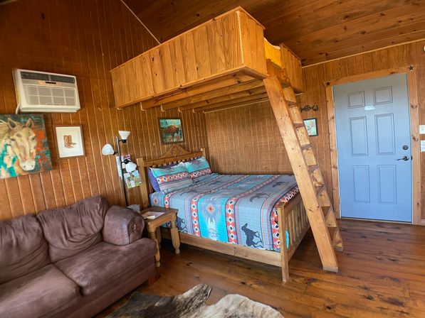 Cozy rustic cabin interior with wood-paneled walls, wooden loft and ladder, queen bed with Southwestern quilt, brown sofa, air conditioner and cowhide rug.