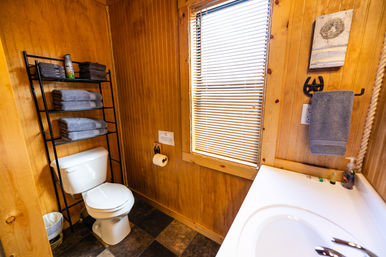 Cozy rustic cabin bathroom with wood-paneled walls, white toilet, black shelving of folded gray towels, window blinds and white sink