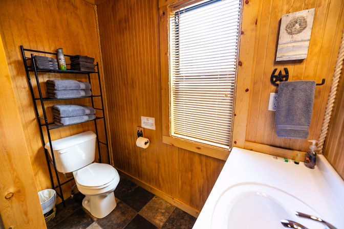 Cozy rustic cabin bathroom with wood-paneled walls, white toilet, black shelving of folded gray towels, window blinds and white sink