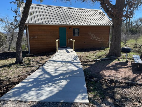 Rustic wooden cabin with silver metal roof and teal front door at the end of a concrete walkway, shaded by oak trees on a sunny hillside with a charcoal kettle grill and picnic bench nearby.