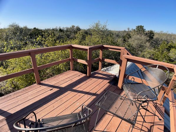 Sunlit wooden deck with railing and metal patio chairs overlooking a green treetop canopy under a clear blue sky.