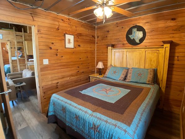 Rustic wood-paneled cabin bedroom with queen bed in a turquoise-and-brown Texas-star quilt, wooden headboard, bedside lamp, ceiling fan, and view into a cozy sitting area.