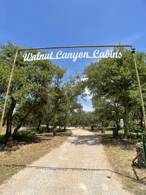 Rustic metal arch with white script sign over a gravel driveway leading into a shaded live-oak grove, sunny blue sky above — entrance to a wooded cabin retreat.