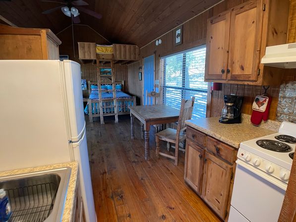 Cozy rustic cabin interior with wood-paneled walls and floors, compact kitchenette (stove, fridge, sink, coffee maker), wooden dining table and chairs, a bed with ladder and overhead storage, and a large sunlit window with blinds.