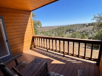 Sunlit wooden cabin porch with rocking chairs and railing overlooking rolling wooded hills and a clear blue sky.