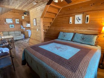Cozy rustic wood-paneled cabin interior with a large star-motif quilted bed, staircase to a loft, compact kitchenette and beige sofa under a ceiling fan, framed bird art and warm lamp light.
