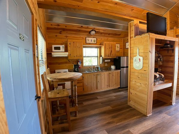 Cozy sunlit rustic cabin kitchenette with knotty pine walls and cabinets, round wooden bistro table with stools, stainless mini-fridge, microwave, sink under window, built-in bunk nook and wall-mounted TV.