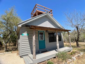 Cozy gray cabin with a teal front door and decorative glass, rustic wooden porch posts, small covered porch and rooftop observation deck set in a wooded rural landscape under a clear blue sky.