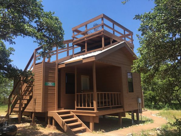 Rustic elevated wooden cabin with front porch, exterior stairs and rooftop deck nestled among oak trees under a clear blue sky