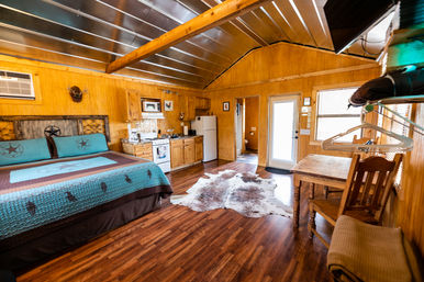 Cozy rustic wood-paneled cabin studio with king bed, turquoise quilt, kitchenette, cowhide rug and dining table under a vaulted metal ceiling