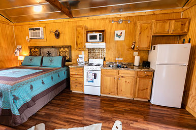 Cozy rustic wooden cabin studio interior with bed topped by turquoise western quilt, compact kitchenette with white refrigerator, stove and microwave, wooden cabinets, sink and hardwood floor.