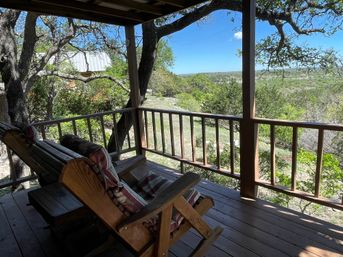 Inviting covered wooden porch with cushioned wood chairs and a swing, shaded by oak branches and overlooking a rolling wooded valley under a clear blue sky.