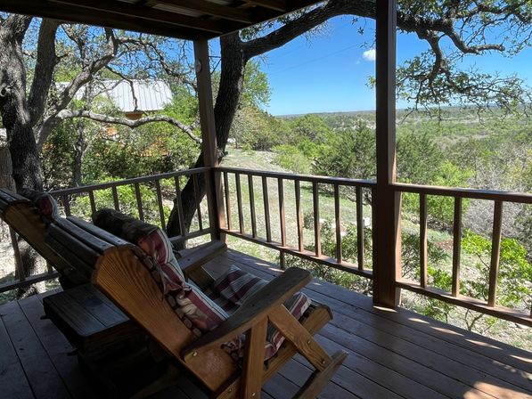 Inviting covered wooden porch with cushioned wood chairs and a swing, shaded by oak branches and overlooking a rolling wooded valley under a clear blue sky.