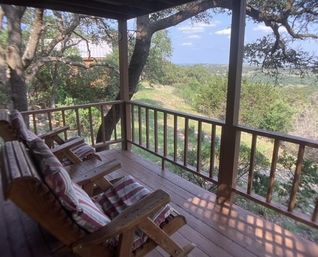 Cozy rustic wooden porch with cushioned lounge chairs overlooking oak-studded hillside and distant valley under a sunny blue sky