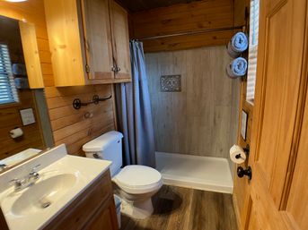 Cozy rustic cabin bathroom with wood paneling, white sink and toilet, walk-in tiled shower with patterned accent, gray curtain, rolled towels and wooden cabinetry.