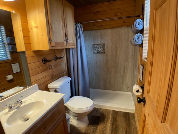 Cozy rustic cabin bathroom with wood paneling, white sink and toilet, walk-in tiled shower with patterned accent, gray curtain, rolled towels and wooden cabinetry.