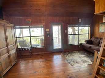 Sunlit rustic wood‑paneled cabin living room with large windows and glass door opening to a tree-lined deck, hardwood floors, cowhide rug, brown sofa and wooden armoire.