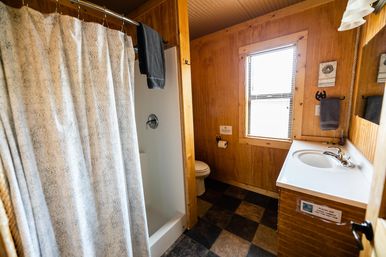 Cozy rustic cabin bathroom with wood-paneled walls, patterned shower curtain, single-sink vanity, toilet, window with blinds, and checkered tile floor.
