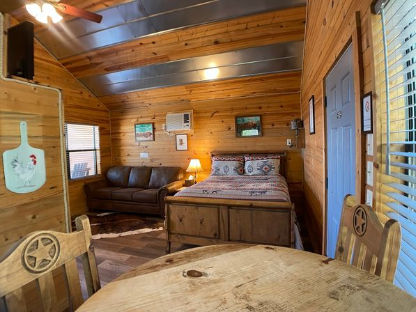 Cozy wood-paneled cabin studio with open-plan bedroom and living area — queen bed with patterned quilt, leather sofa, rustic round dining table, ceiling fan and window AC.