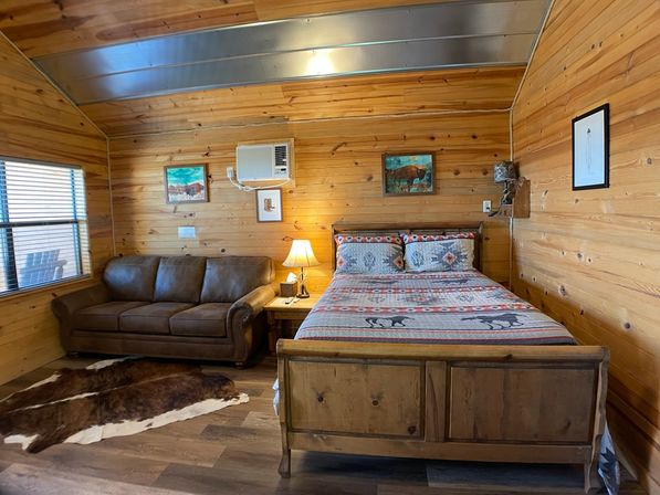 Cozy rustic wood-paneled cabin bedroom with patterned quilted queen bed, leather sofa, cowhide rug, nightstand lamp, wall art and window air conditioner.
