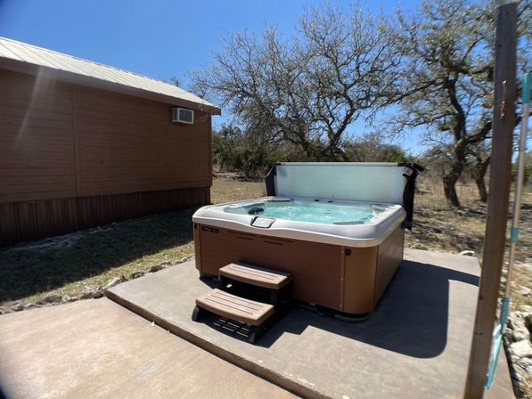 Inviting outdoor hot tub with wooden steps on a concrete patio next to a cabin, bubbling under a clear blue sky with sparse trees in a rural backyard.