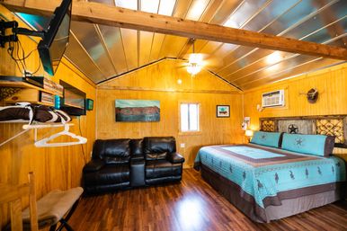 Cozy rustic cabin bedroom with wood-paneled walls and metal vaulted ceiling, turquoise king bed, black leather loveseat, hardwood floors, wall air conditioner